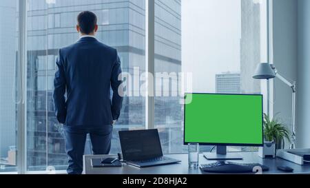 Confident Businessman in a Suit Contemplating Business Deal in His Office, Looking out of the Window. Desctop PC has Green Screen Mock-up Template Stock Photo