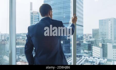 Confident Businessman in a Suit Looking out of the Window Thoughtfully. Stylish Modern Business Office with Personal Computer and Big City View. Stock Photo