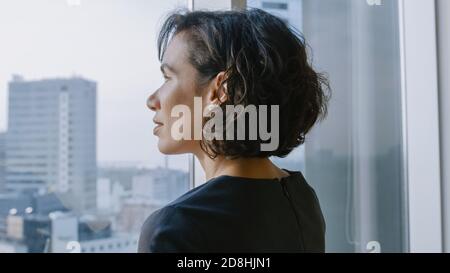 Close-up Portrait of Successful Businesswoman in the Striking Black Dress Standing in Her Office and Looking out of the Window Thoughtfully. Modern Stock Photo