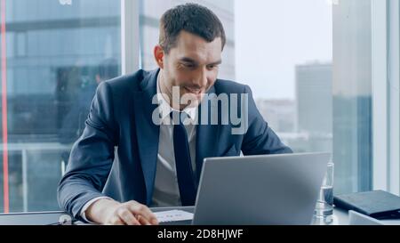 Successful Businessman Sitting at His Desk, Using His Laptop. In the Window Big City Business District View. Stock Photo