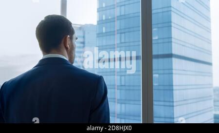Medium Shot of Successful Businessman wearing a Suit Standing in His Office, Contemplating Next Big Business Deal, Looking out of the Window. Big City Stock Photo