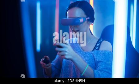 Beautiful Pro Gamer Girl Wearing Virtual Reality Headset Plays in Online Video Game, Gesturing Using Controllers. Cool Retro Neon Colors in the Room. Stock Photo
