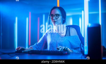 Beautiful Professional Gamer Girl Playing and Streaming Online Video Game on Her Personal Computer. Casual Cute Geek wearing Glasses. Cyber e-Sport Stock Photo