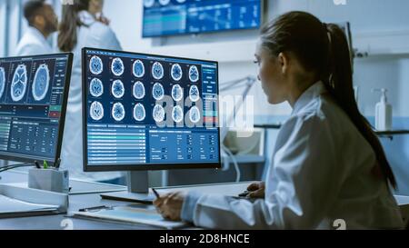 Female Medical Research Scientist Working with Brain Scans on Her Personal Computer, Writing Down Data in a Clipboard. Modern Laboratory Working on Stock Photo