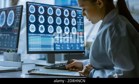 Female Medical Research Scientist Working with Brain Scans on Her Personal Computer, Writing Down Data in a Clipboard. Modern Laboratory Working on Stock Photo