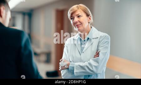 Portrait Shot of a Senior Female Doctor Standing in the Hospital Hallway and Having Professional Discussion. Modern Hospital with Patients and Medical Stock Photo