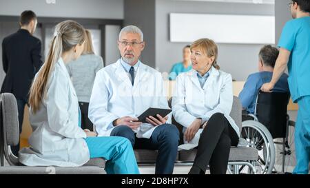 In the Lobby of the Hospital Three Professional Doctor Have Discussion while Sitting. They Use Tablet Computer. Busy Modern Hospital with Best Stock Photo