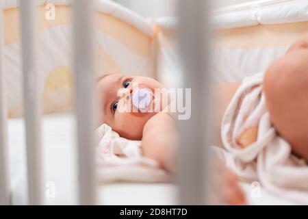 Portrait of a cute caucasian baby with a pacifier in his mouth, who is lying in a cradle. View through the bars of the cradle. Stock Photo