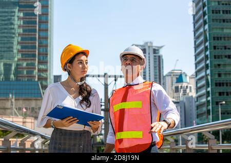 Female industrial engineer wearing a white helmet while standing in a construction site with businessman talking on working plan, Engineer and archite Stock Photo