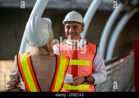 Female industrial engineer wearing a white helmet while standing in a construction site with businessman talking on working plan, Engineer and archite Stock Photo