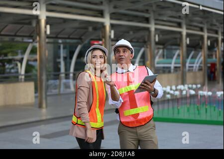 Female industrial engineer wearing a white helmet while standing in a construction site with businessman talking on working plan, Engineer and archite Stock Photo
