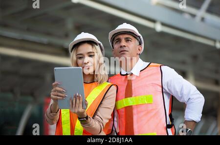 Female industrial engineer wearing a white helmet while standing in a construction site with businessman talking on working plan, Engineer and archite Stock Photo