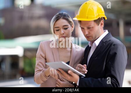 Female industrial engineer wearing a white helmet while standing in a construction site with businessman talking on working plan, Engineer and archite Stock Photo