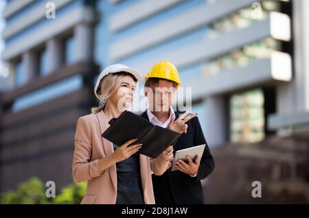 Female industrial engineer wearing a white helmet while standing in a construction site with businessman talking on working plan, Engineer and archite Stock Photo