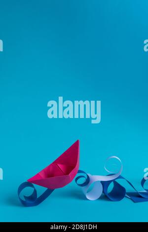 pink paper boat navigating on paper waves on bright blue background. Stock Photo