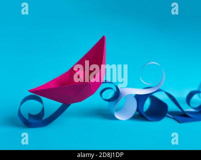 pink paper boat navigating on paper waves on bright blue background. Stock Photo