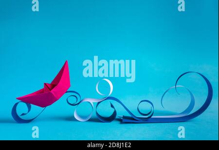 pink paper boat navigating on paper waves on bright blue background. Stock Photo