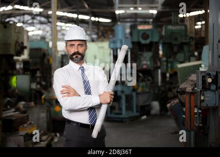 The bearded hipster director with engineer hat standing against machine in production in factory Stock Photo