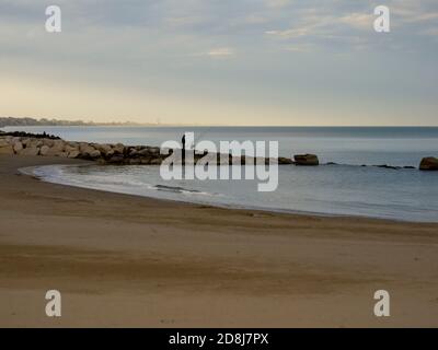 Person fishing from rocks in a sandy bay in Italy Stock Photo