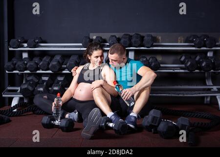 Sport family pregnancy in urban cross training gym. Sport and healthy lifestyle during pregnancy. Young couple hugging on gym floor after working out Stock Photo