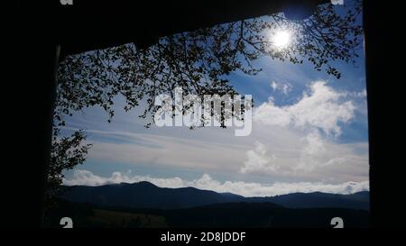 View through a window from a wooden house in the forest with the sun in the sky surrounded by clouds and with the Black Forest in the background Stock Photo