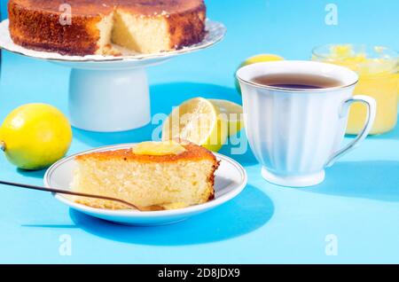 Piece of Homemade freshly baked sponge cake on plate, Lemon Kurd in a glass jar with spoon, whole and sliced fresh lemons on plate over blue backgroun Stock Photo