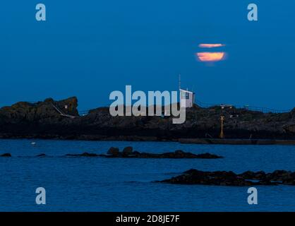 North Berwick, East Lothian, Scotland, United Kingdom, 30th October 2020. UK Weather: the rise of a Hunter moon with the harbourmaster's cabin on the rocky shore seen from across West Bay. This is the second full moon in the month of October, making it also a Blue Moon Stock Photo
