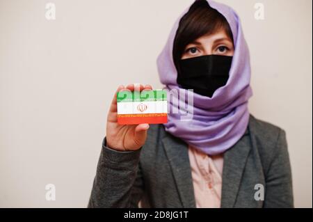 Portrait of young muslim woman wearing formal wear, protect face mask and hijab head scarf, hold Iran flag card against isolated background. Coronavir Stock Photo