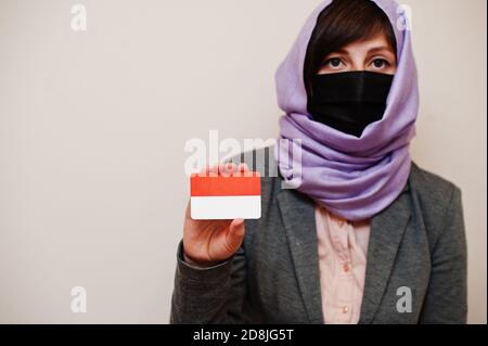 Portrait of young muslim woman wearing formal wear, protect face mask and hijab head scarf, hold Indonesia flag card against isolated background. Coro Stock Photo