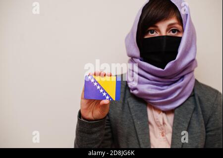 Portrait of young muslim woman wearing formal wear, protect face mask and hijab head scarf, hold Bosnia and Herzegovina flag card against isolated bac Stock Photo