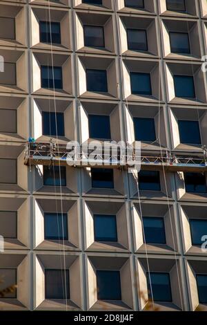 Two man are seen on an elevated platform hanging on the side of a skyscraper. These workers wearing belts and helmets are cleaning the windows at a hi Stock Photo