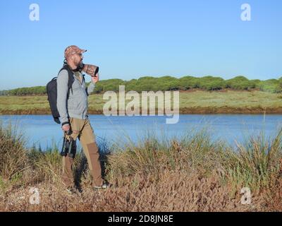 young hipster man with a beard in the field carrying a photographic equipment with a reflex camera and a telephoto lens, binoculars and backpack Stock Photo