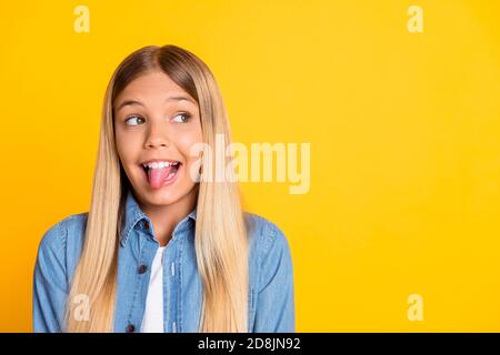 Photo portrait of funny playful girl with long blonde hair showing tongue wearing denim shirt isolated on vivid yellow background with blank space Stock Photo