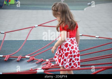 the girl on the playground beautiful cute pretty girl in a dress with red hearts ingest near a children's climbing network Stock Photo