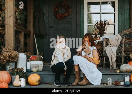 children a boy and a girl with their poodle dog in protective masks on the porch of the backyard decorated with pumpkins in autumn, the concept of the Stock Photo