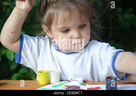 A girl of 2 3 years old sits at a table and draws with colorful paints. Children's creativity. Children's hobbies. Stock Photo