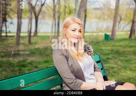 Thoughtful. Portrait of a serious woman looking away to side sitting on a bench in a city park green trees background. Beautiful middle aged lady in g Stock Photo