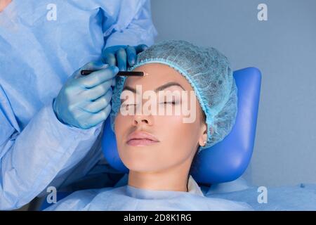 Doctor preparing for forehead lift surgery procedure about to draw lift lines with marker pencil on patient woman face in a clinic surgery room, O.R b Stock Photo