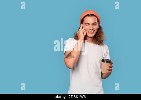 Freestyle. Curly-haired guy wearing hat standing isolated on blue with cup of coffee talking on smartphone smiling happy Stock Photo