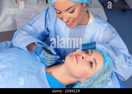 Anti-aging treatment and face lift. Beautiful doctor woman is about to draw lift lines on patient neck with pencil for lifting procedure. Two people i Stock Photo