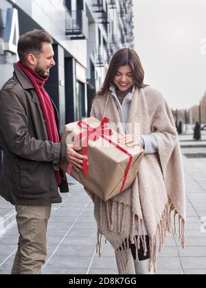 Man giving a Christmas present to his girlfriend. Modest embarrassed woman accepts a large wrapped holiday gift on the street. Stock Photo