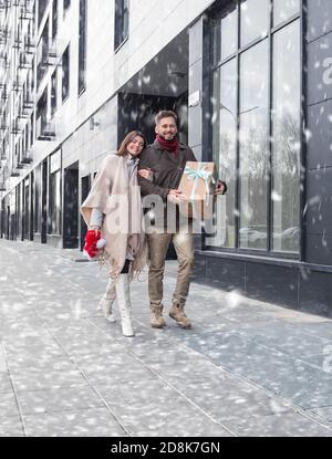 Happy laughing winter young couple walking in the street with Christmas gift present. Woman laid her head on her husband. Young couple holding big box Stock Photo