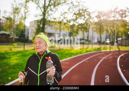 Old woman walking with nordic walk sticks on running track, rubber treadmill and stopped to quench thirst, drink water from a flask. Active female Stock Photo