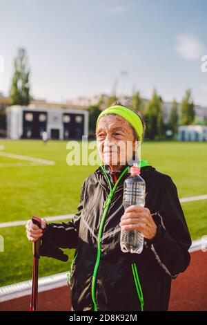 Old woman walking with nordic walk sticks on running track, rubber treadmill and stopped to quench thirst, drink water from a flask. Active female Stock Photo