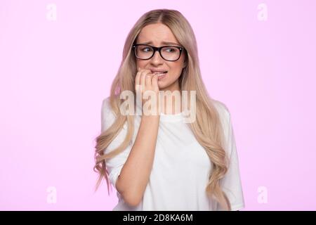 Worried young woman looks nervous, bites finger, isolated. Female in eyeglasses feels frustrated due to an awkward situation, has a problem, has no id Stock Photo