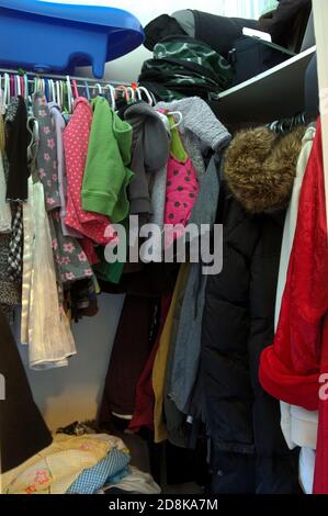 Small, cramped closet with many clothes hanging on hangers and things on shelves Stock Photo