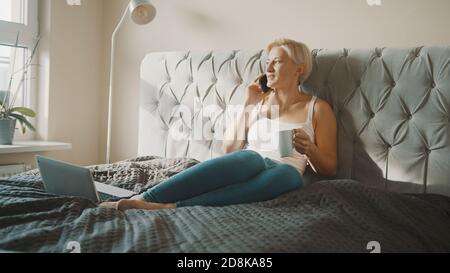 Young blond woman having a phone call while drinking coffee or tea in her bedroom. High quality photo Stock Photo