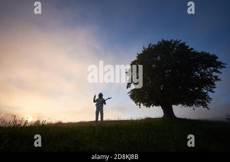 Two figures on purple sky background, of man astronaut in space suit holding guitar and showing peace sign and a big old tree on the right Stock Photo