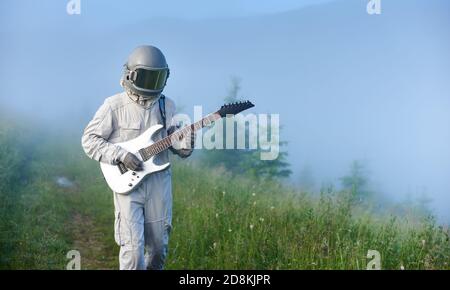 Astronaut in space suit walking on grassy path and playing on guitar. Cosmonaut guitarist with musical instrument strolling down foggy meadow with green grass. Concept of music, astronautics, nature. Stock Photo