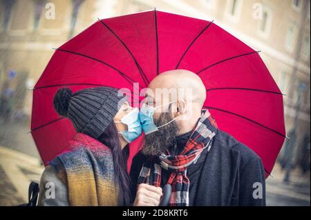 Romantic happy couple kissing with face mask in the street standing under an umbrella in the rain in the covid-19 time - New normal love concept - Col Stock Photo